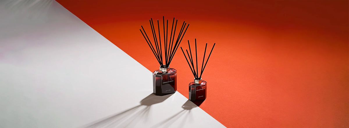 Two dark glass reed diffusers with black sticks on white and orange surface