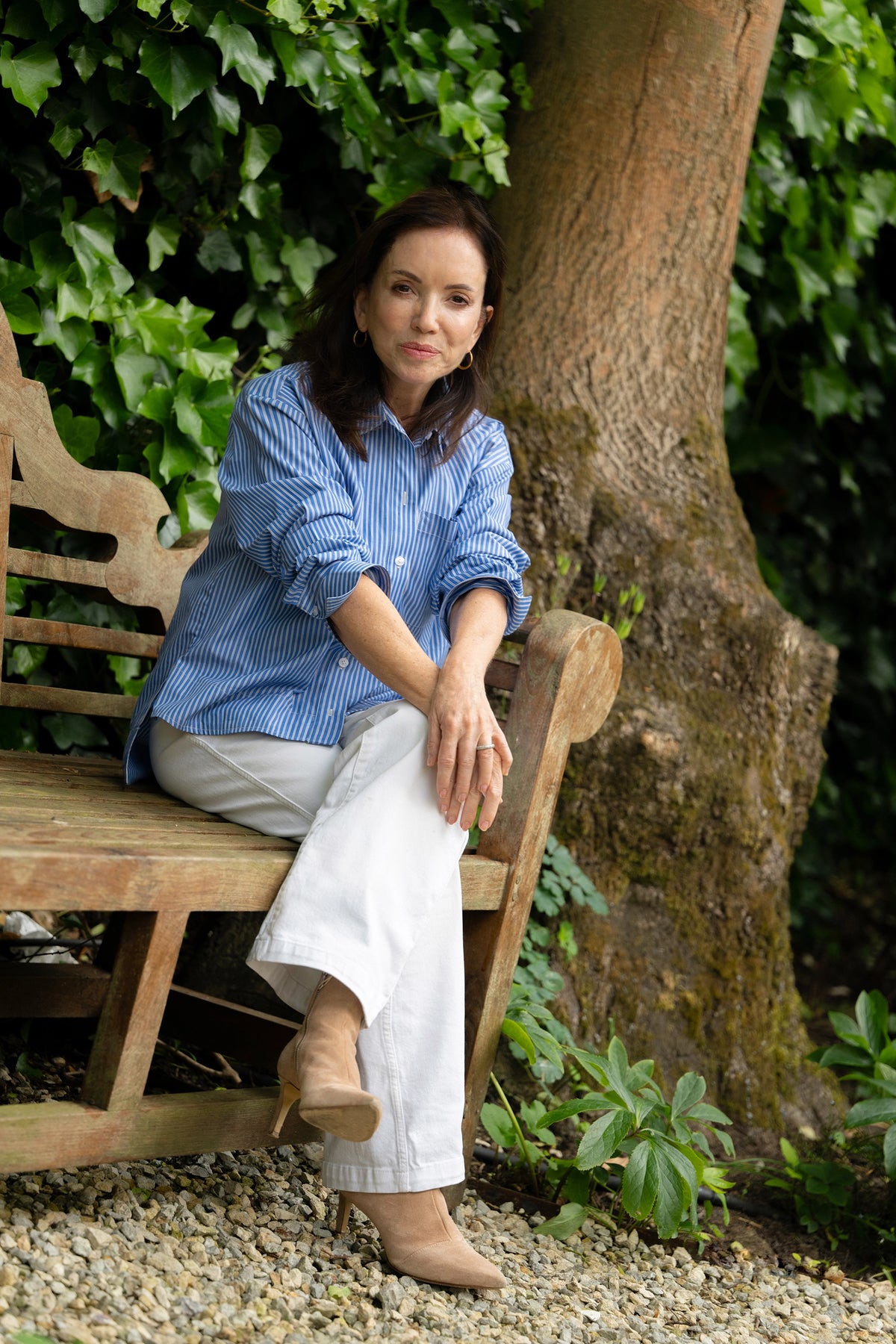 Woman wearing blue striped shirt, white trousers, and beige heeled boots sitting on wooden bench in garden